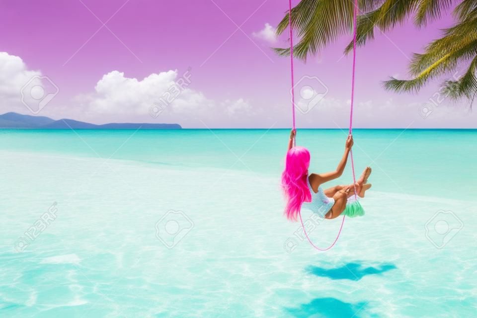 Back view of girl with pink hear having fun on swing hanging on tree at tropical beach with white sand. Luxury vacation on paradise island.