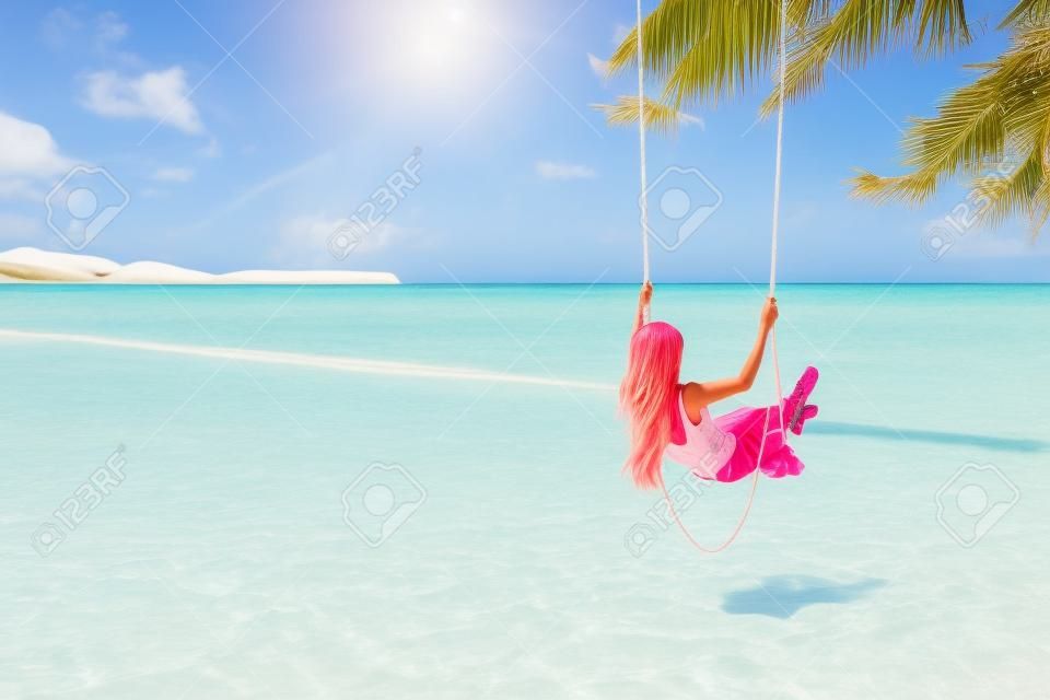 Il punto di vista posteriore della ragazza con il rosa sente divertirsi sull'oscillazione che appende sull'albero alla spiaggia tropicale con sabbia bianca. Vacanza di lusso sull'isola paradisiaca.