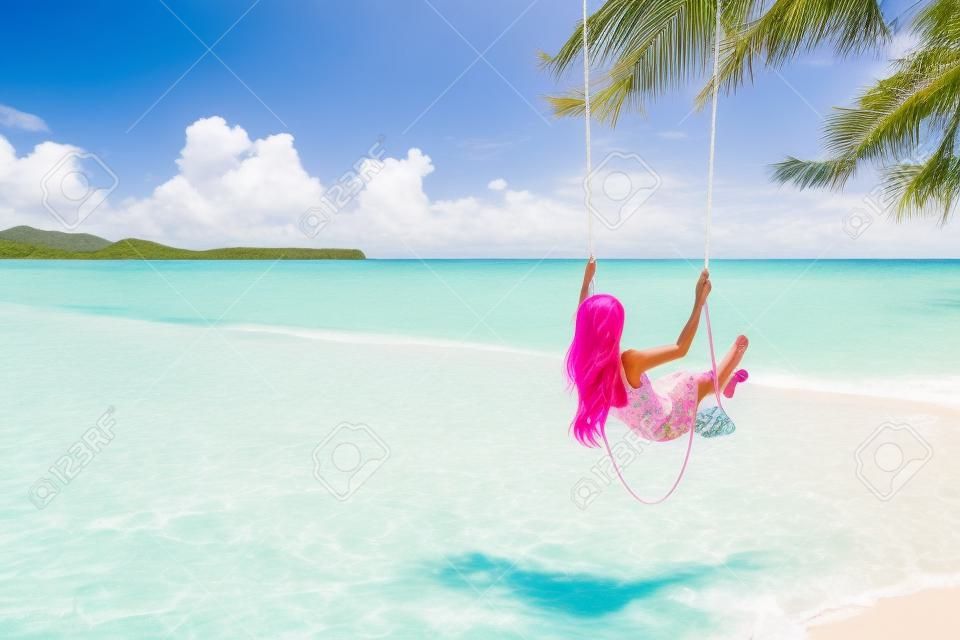 Back view of girl with pink hear having fun on swing hanging on tree at tropical beach with white sand. Luxury vacation on paradise island.