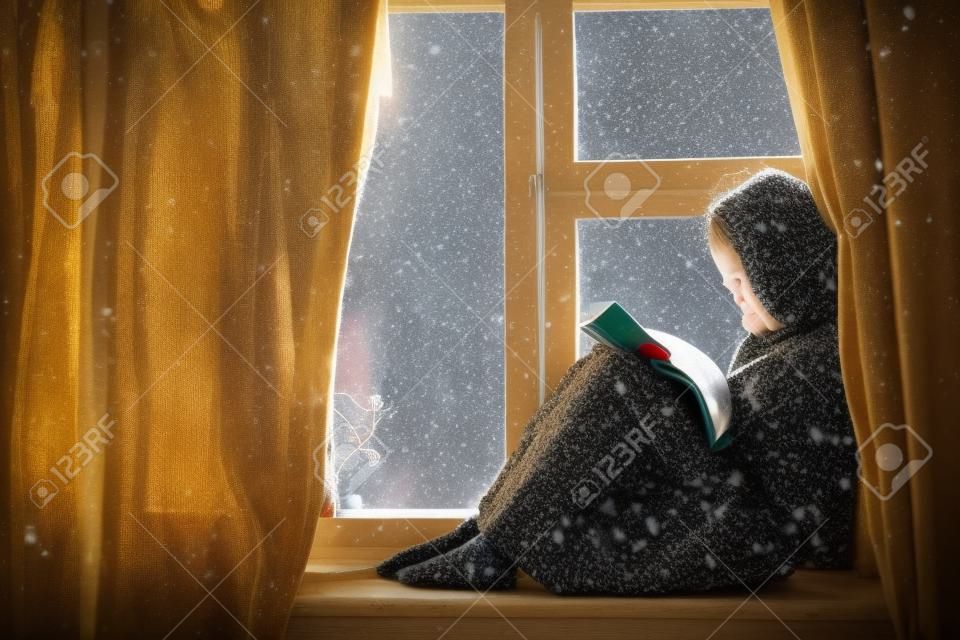 Enfant pré-ado en pull de laine chaud assis sur le rebord de la fenêtre et lisant un livre. Week-ends d'hiver dans la vieille maison en rondins. Temps froid et neigeux. Concept chaleureux et accueillant.