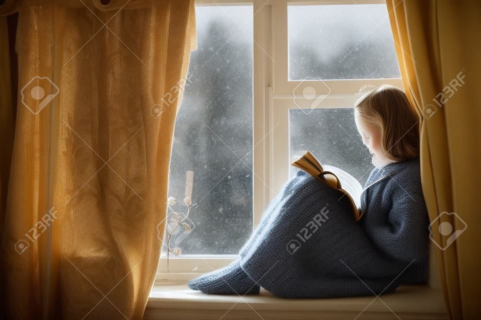 Pre Teen Kind in warmen Wollpullover sitzt auf der Fensterbank und liest ein Buch. Winterwochenenden im alten Blockhaus. Kaltes Schneewetter. Gemütliches Wohnkonzept.