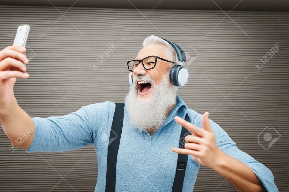 Älterer verrückter Mann, der sich selbst filmt, während er Musik mit Kopfhörern hört - Hipster-Typ, der Spaß mit mobilen Smartphone-Playlist-Apps hat - Konzept von Glück, Technologie und älteren Menschen