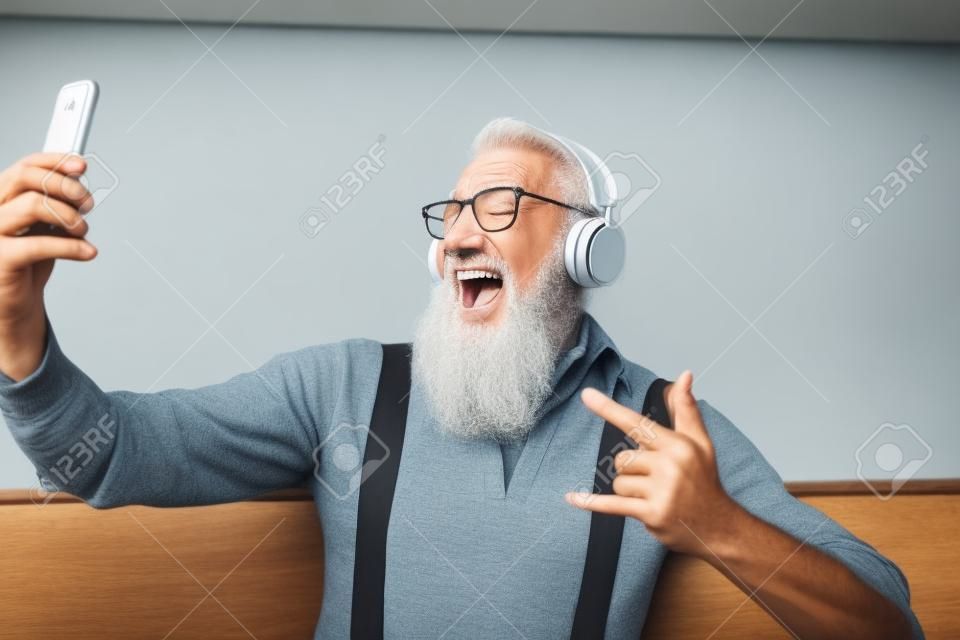 Älterer verrückter Mann, der sich selbst filmt, während er Musik mit Kopfhörern hört - Hipster-Typ, der Spaß mit mobilen Smartphone-Playlist-Apps hat - Konzept von Glück, Technologie und älteren Menschen