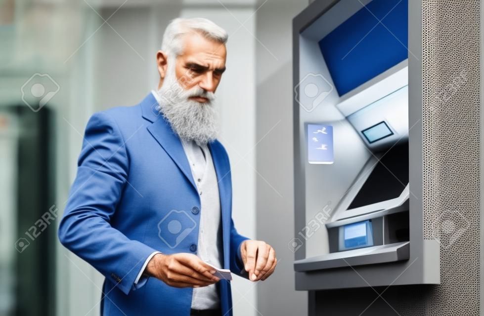 Starszy mężczyzna wypłaca pieniądze z bankomatu z kartą debetową - brodaty hipster dojrzały mężczyzna dokonujący płatności kartą kredytową w bankomacie - koncepcja biznesu, konta bankowego i starszych ludzi stylu życia