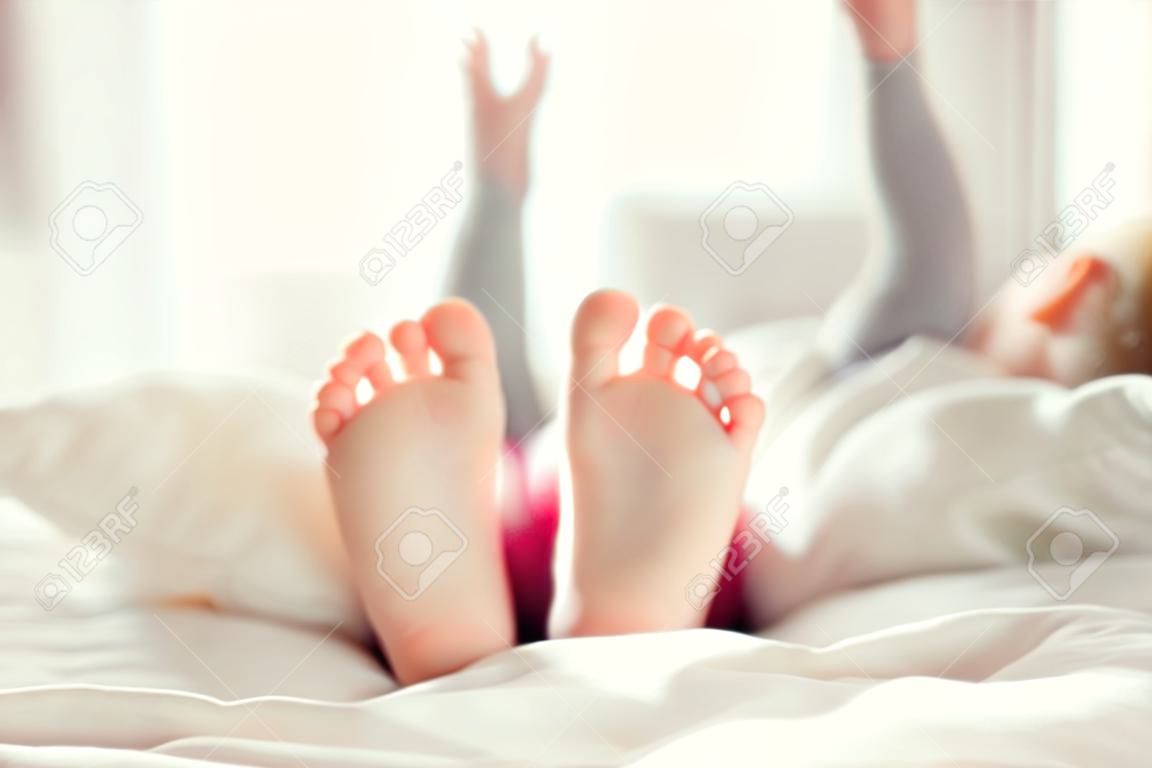 Pies de niño en la manta blanca en cama. Niña adorable que despierta en su cama. Mañana. Retrato de los pies de los niños en el dormitorio