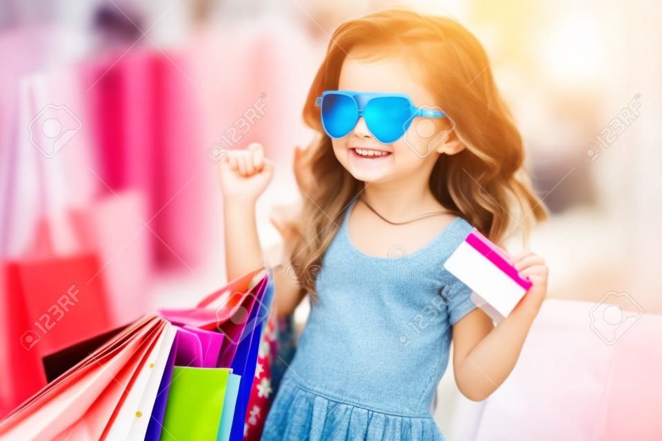 Schönes, glückliches, lächelndes kleines Mädchen mit Sonnenbrille hält Einkaufstaschen in der Nähe des Einkaufszentrums im Freien. Lifestyle-Konzept. Kleines Shopaholic-Mädchen. Mädchen mit Taschen in den Händen. Die Freude am Einkaufen.