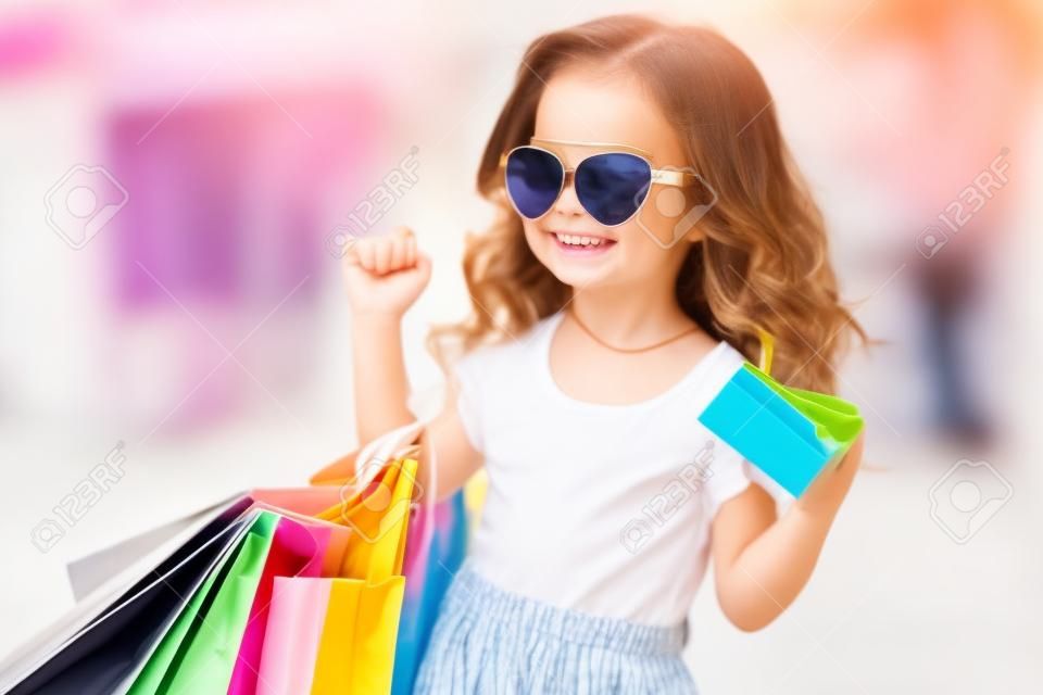 サングラスをかけた美しい幸せな笑顔の小さな女の子の子供は、屋外のショッピングモールの近くで買い物袋を持っています。ライフスタイルのコンセプト。買い物中毒の女の子。手にバッグを持つ女の子。買い物の喜び。