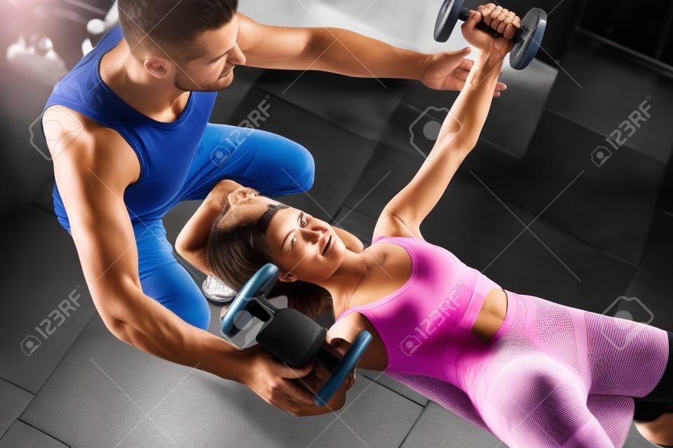 Im Fitnessstudio unterstützt ein männlicher Personal Trainer eine Frau beim Heben einer Hantel. Männlicher Personaltrainer zur sportlichen Unterstützung, der Frau beim Gewichtheben unterstützt