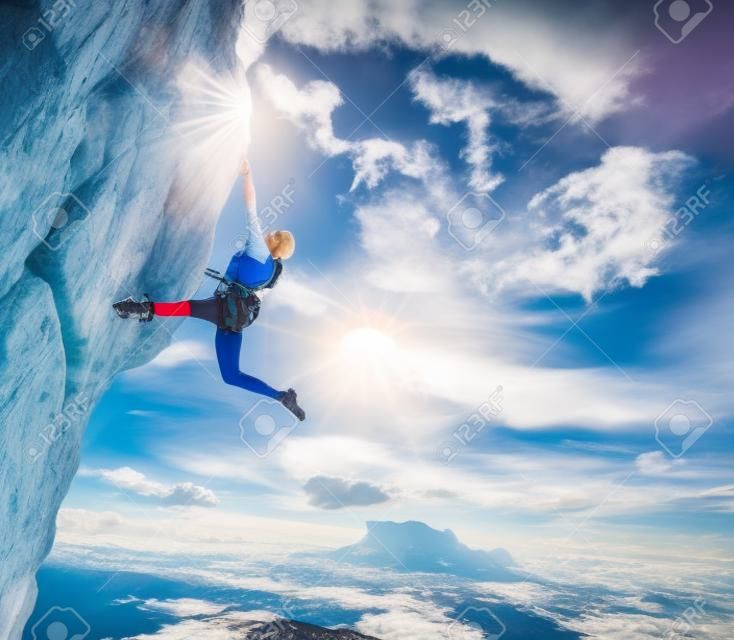 Atleta fêmea elegante que pendura no topo do pico perigoso equipado com o arnês da corda da engrenagem céu azul nuvens fantásticas no fundo e raios de sol que brilham de cima