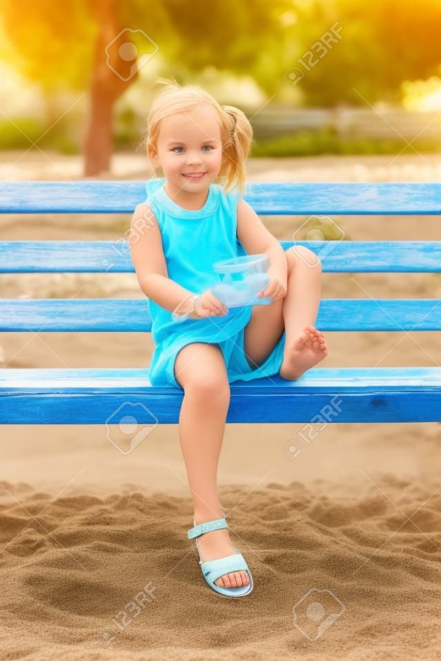 Beautiful little girl sitting on an old bench shakes the sand out of the flip-flops
