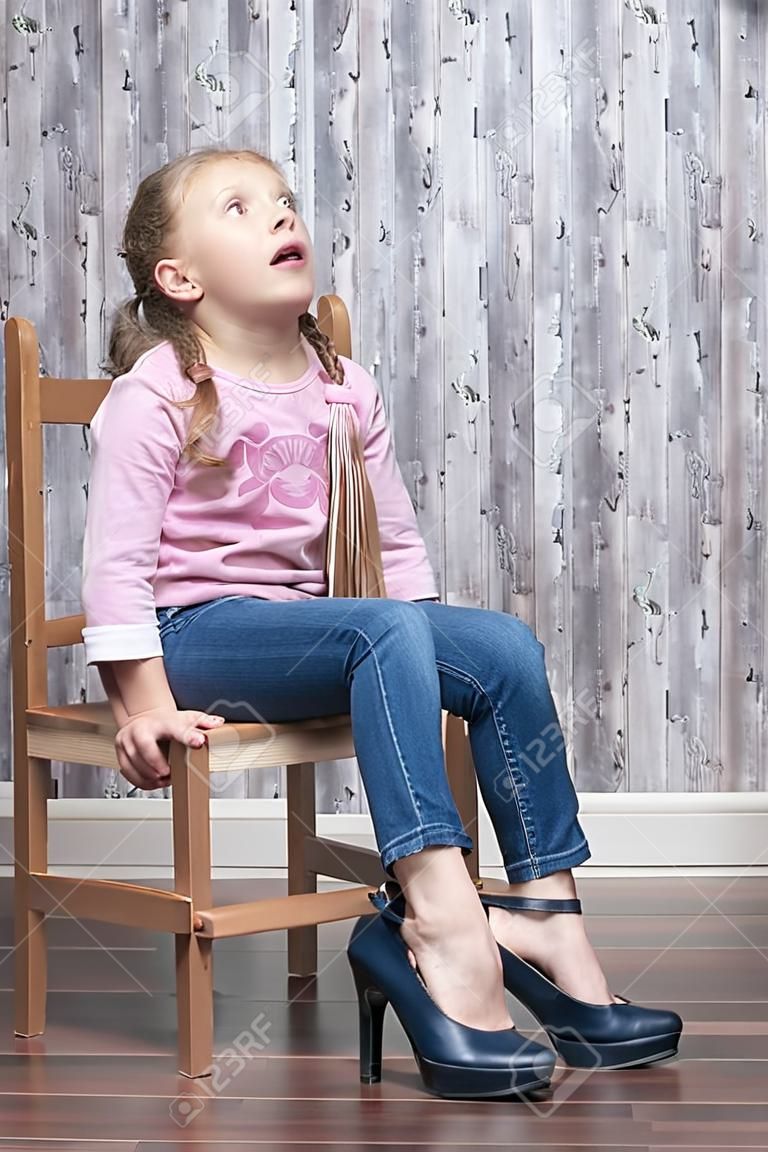young girl sitting on a wooden chair looks frustrated up trying on her mother's high-heeled shoes