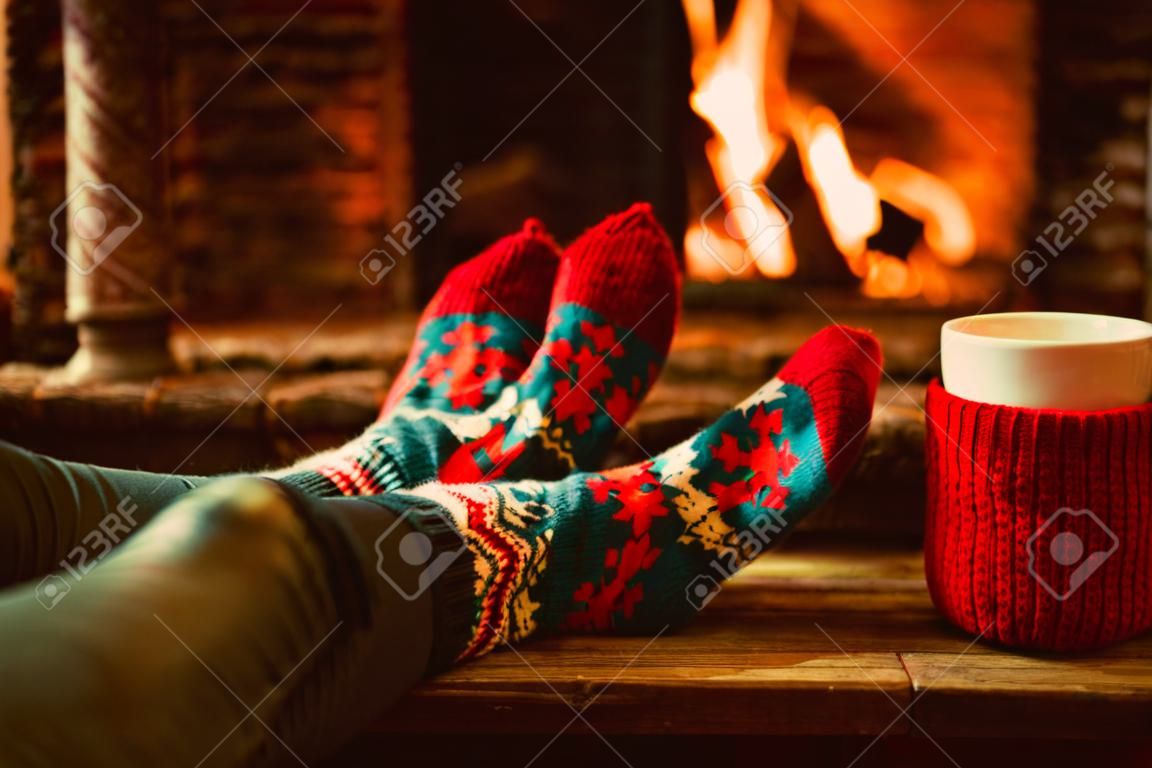 Pies en calcetines de lana junto a la chimenea de la Navidad. La mujer se relaja por el fuego caliente con una taza de bebida caliente y calentar sus pies en calcetines de lana. Cerca de los pies. Invierno y vacaciones de Navidad concepto.