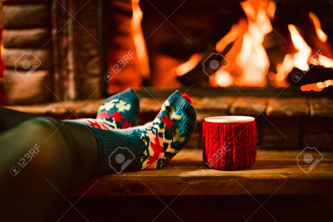 Füße in Wollsocken von der Weihnachts Kamin. Frau entspannt sich durch warmes Feuer mit einer Tasse heißes Getränk und Aufwärmen ihre Füße in Wollsocken. Schließen Sie oben auf die Füße. Winter-und Weihnachtsferien Konzept.