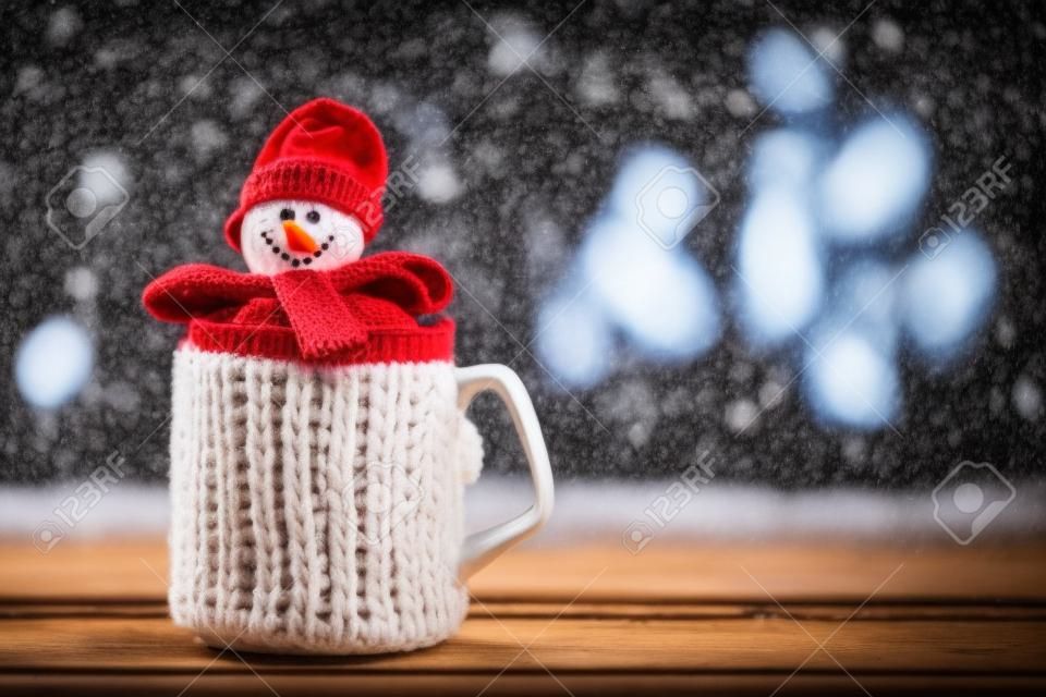 Cup of hot drink in front of warm fireplace. Holiday Christmas concept. Mug in red knitted mitten, decorated with snowman toy, standing near fireside. Cozy relaxed magical atmosphere in a chalet.