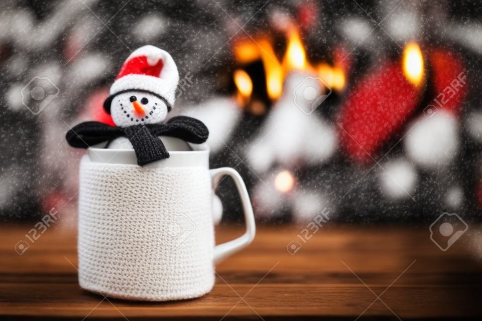 Cup of hot drink in front of warm fireplace. Holiday Christmas concept. Mug in red knitted mitten, decorated with snowman toy, standing near fireside. Cozy relaxed magical atmosphere in a chalet.