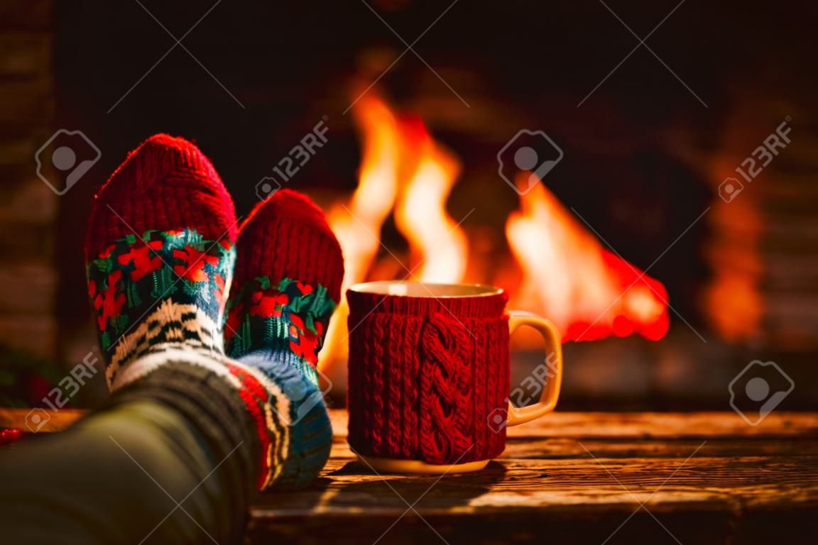 Pies en calcetines de lana junto a la chimenea de la Navidad. La mujer se relaja por el fuego caliente con una taza de bebida caliente y calentar sus pies en calcetines de lana. Cerca de los pies. Invierno y vacaciones de Navidad concepto.