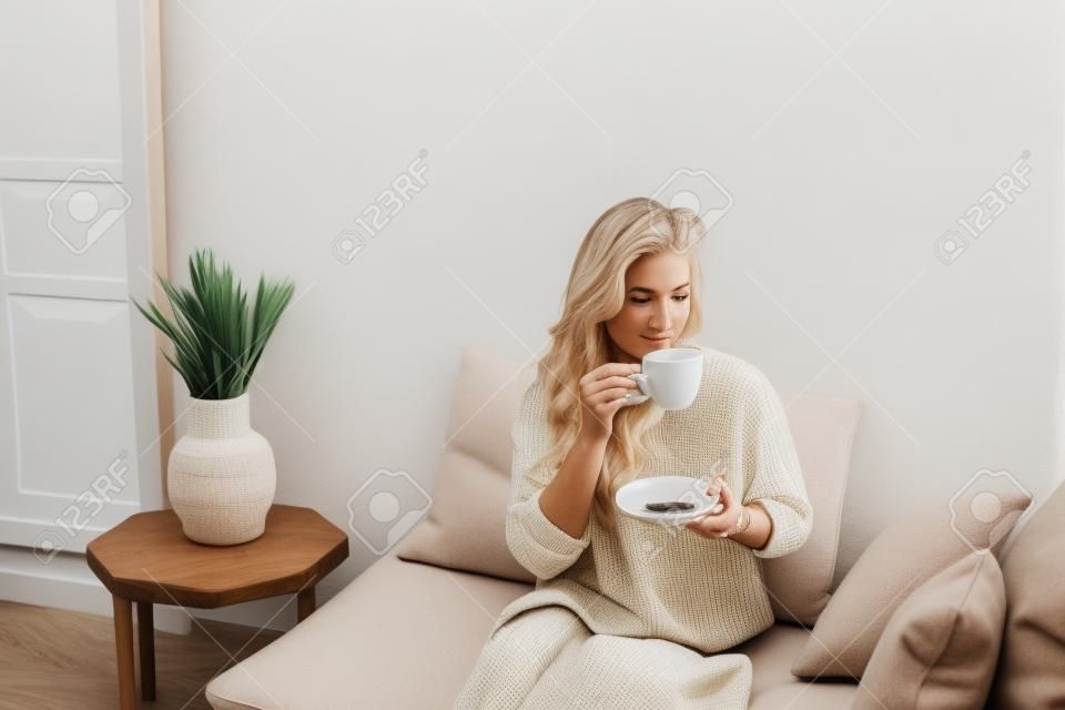 Een jonge blonde vrouw in casual comfortabele kleding zit op een witte bank thuis en drinkt koffie. scandinavian interieur, minimalisme