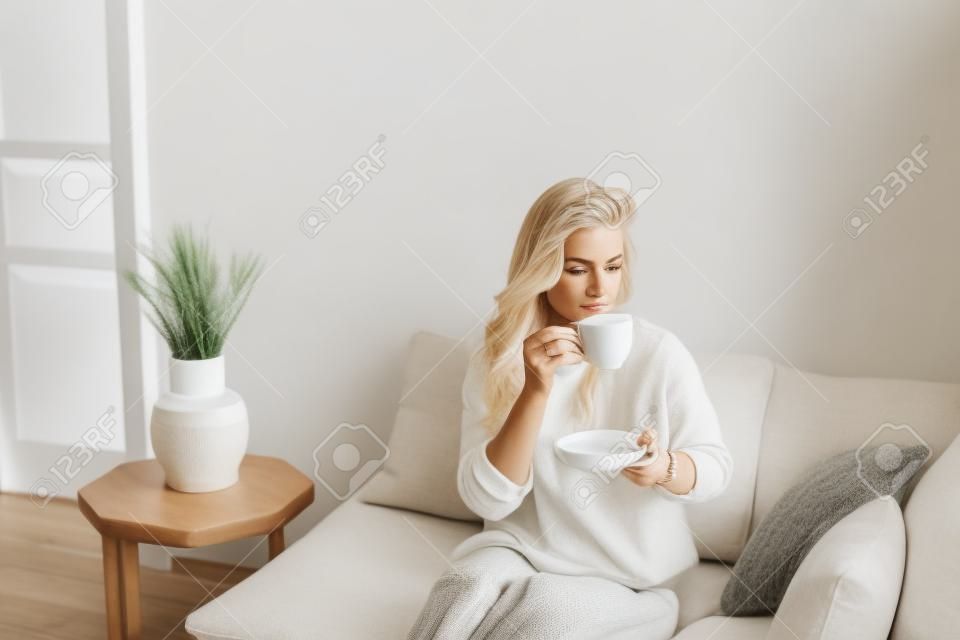 Młoda blondynka w swobodnych wygodnych ubraniach siedzi na białej kanapie w domu i pije kawę. skandynawskie wnętrze, minimalizm