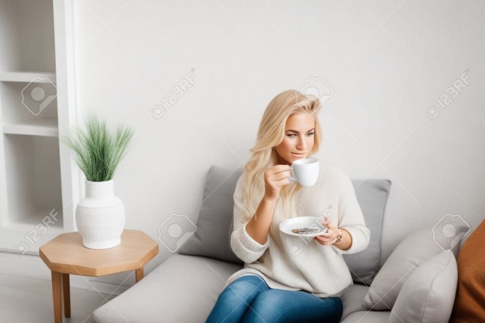 Młoda blondynka w swobodnych wygodnych ubraniach siedzi na białej kanapie w domu i pije kawę. skandynawskie wnętrze, minimalizm