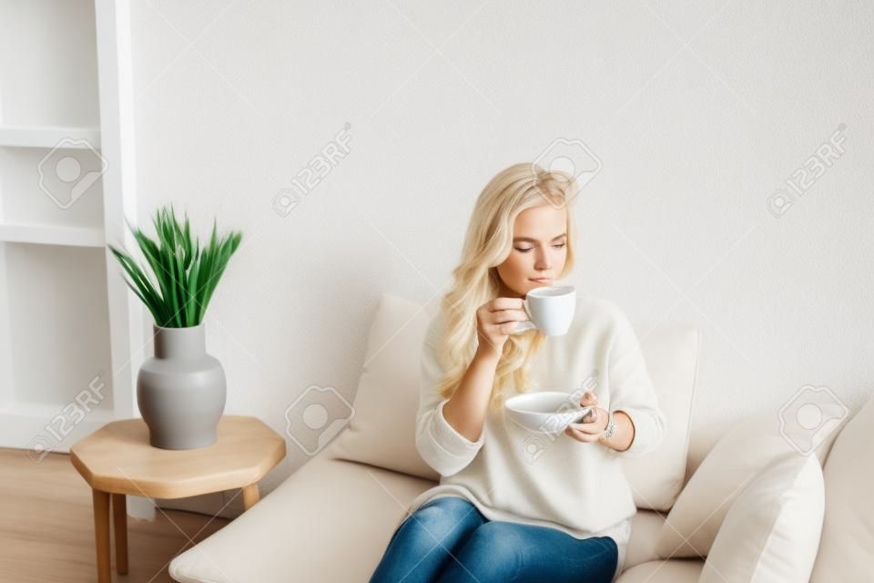 Una joven rubia con ropa informal y cómoda se sienta en un sofá blanco en casa y bebe café. interior escandinavo, minimalismo