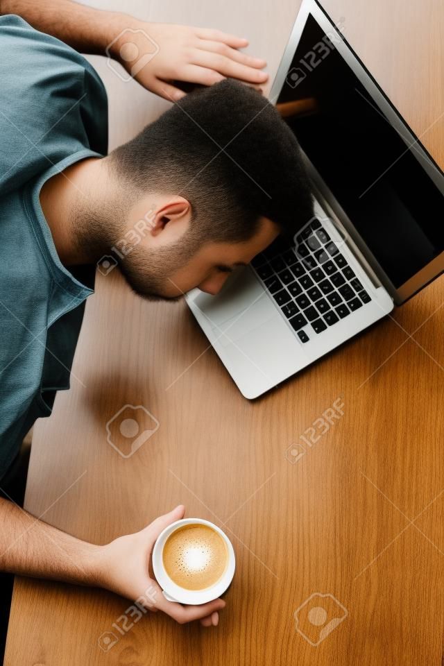 Vertikales Bild des Kerls, der am Tisch sitzt. Er hält eine Tasse Kaffee. Er hat seinen Kopf über den Laptop gelegt. Junger Mann schläft. Er ist müde. Schnittansicht