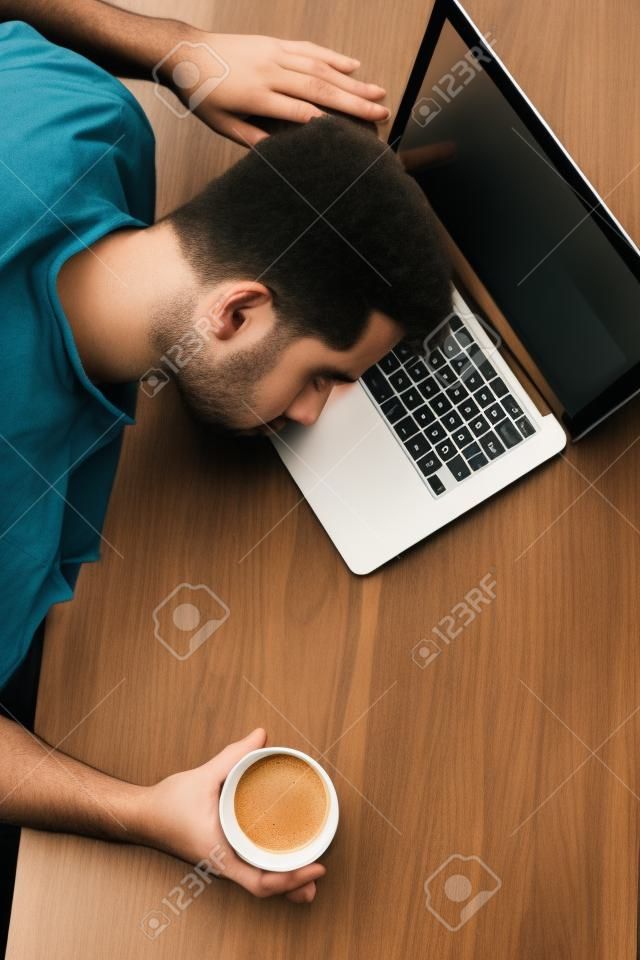 Vertikales Bild des Kerls, der am Tisch sitzt. Er hält eine Tasse Kaffee. Er hat seinen Kopf über den Laptop gelegt. Junger Mann schläft. Er ist müde. Schnittansicht