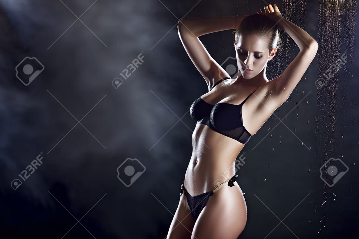 Beautiful wet blonde tall slim girl wearing a black lingerie posing in rain water drops in a studio on black background in a theatrical smoke. Smooth healthy wet skin. Copy space.
