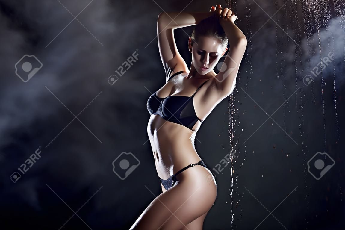 Beautiful wet blonde tall slim girl wearing a black lingerie posing in rain water drops in a studio on black background in a theatrical smoke. Smooth healthy wet skin. Copy space.