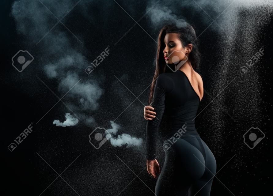 Schönes langbeiniges und beutetes athletisches Fitness-Mädchenmodell, das einen schwarzen Körper trägt, mit nasser fettiger Haut, die unter Wassertropfen im Theaterrauch auf schwarzem Hintergrund posiert. Platz kopieren.