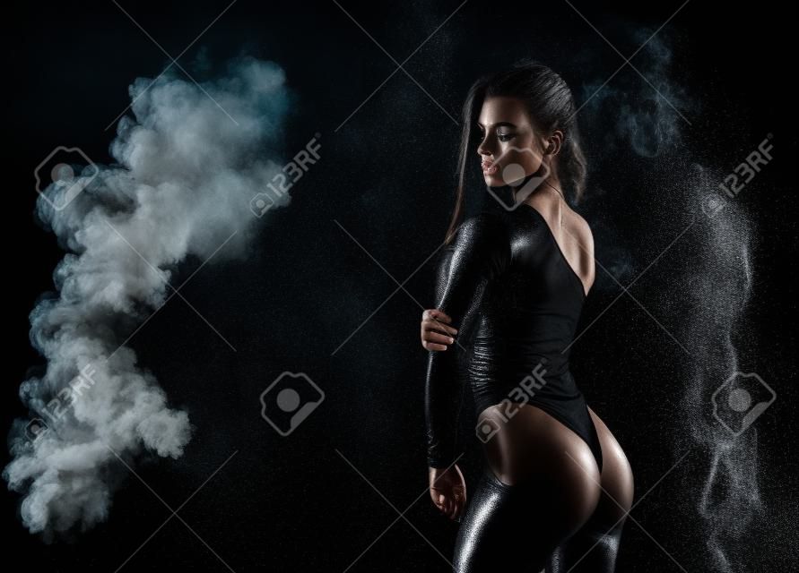 Beautiful leggy and booty athletic fitness girl model, wearing a black body, with wet oily skin, posing under water drops in theatrical smoke on a black background. Copy space.
