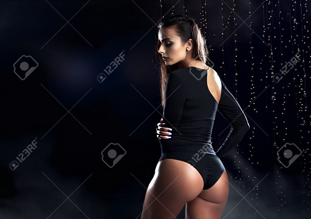 Modello di ragazza bella leggy e bottino atletico fitness, che indossa un corpo nero, con la pelle grassa bagnata, in posa sotto le gocce d'acqua in fumo teatrale su sfondo nero. Copia spazio.