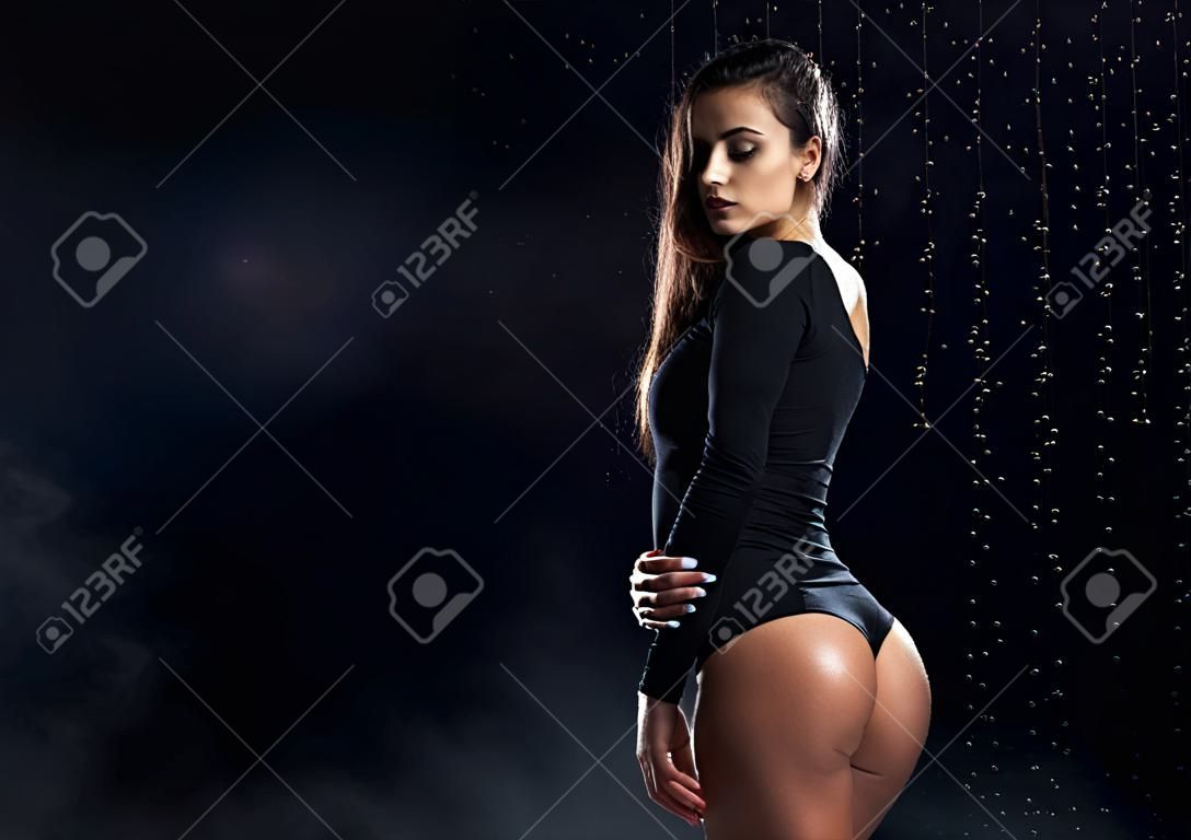Beautiful leggy and booty athletic fitness girl model, wearing a black body, with wet oily skin, posing under water drops in theatrical smoke on a black background. Copy space.