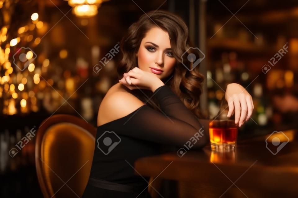Schoonheid jonge brunette vrouw zitten aan de bar met glas whisky in luxe interieur