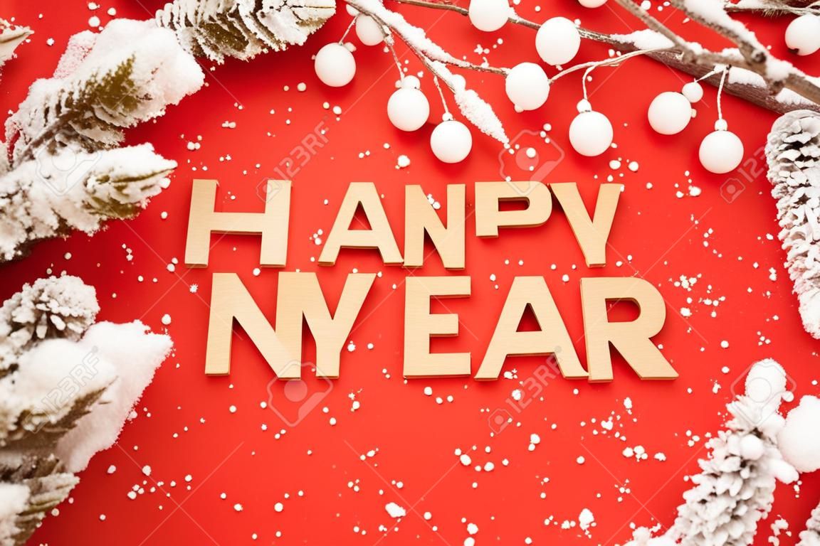 Celebrating Happy New Year. Wooden letters in a New Years cap on a red background with green spruce branches and snow. Festive decoration or postcard concept. Top view and flatlay. Copy space
