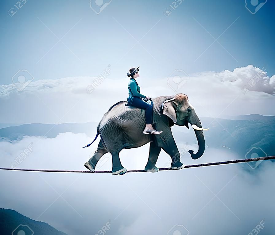 Young woman riding an elephant on a rope at high altitude, above clouds and mountains.