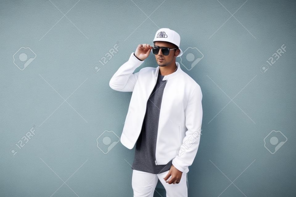 Hombre guapo de moda en una elegante chaqueta blanca con una gorra de  béisbol en gafas de sol posando cerca de la pared