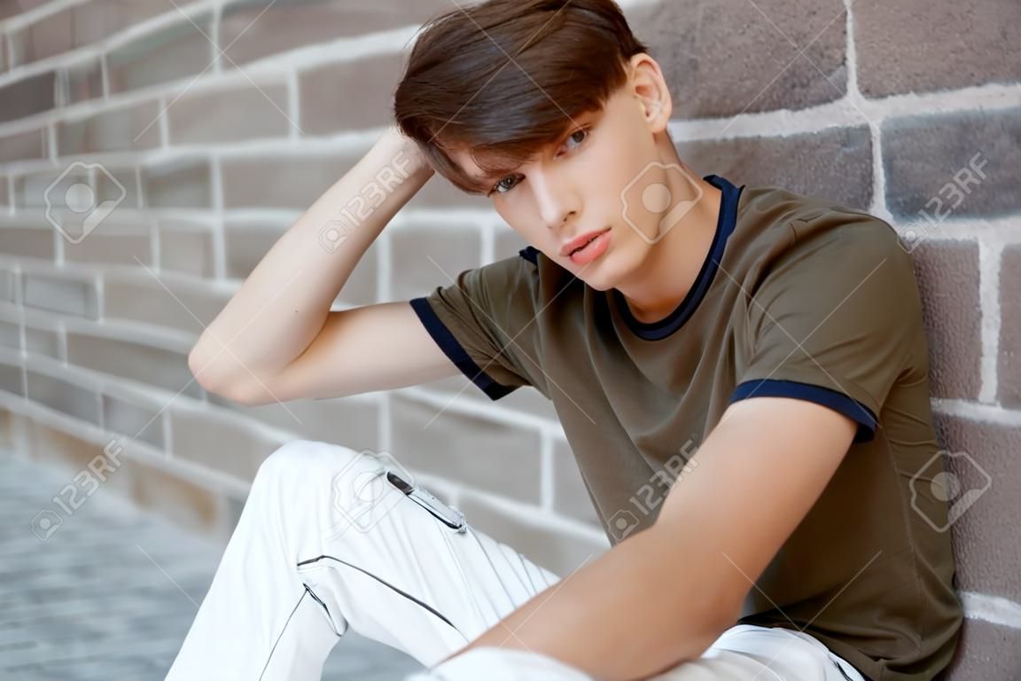 Europäischer junger Mann in einem modischen grünen T-Shirt in stylischen weißen Hosen mit modischer Frisur entspannt sich in der Nähe einer alten Backsteinmauer. Netter moderner Model-Typ ruht sich aus.