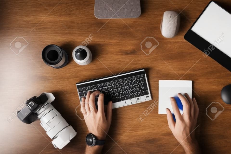 Designer is sitting at workplace and typing on the keyboard. Top view of men's hands and on a table with a photo camera with lenses for camera with a graphic tablet with a mouse. Photographer works.
