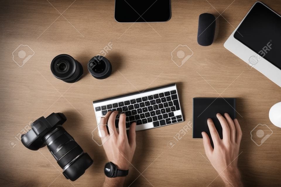 Designer is sitting at workplace and typing on the keyboard. Top view of men's hands and on a table with a photo camera with lenses for camera with a graphic tablet with a mouse. Photographer works.