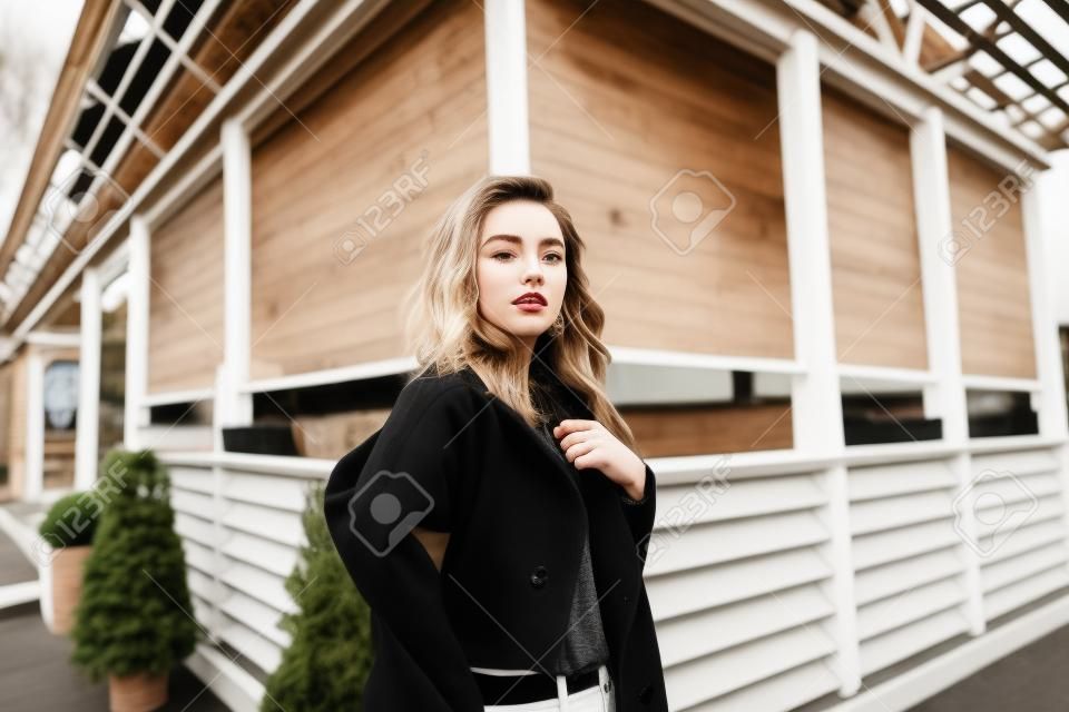 Belle jeune femme urbaine dans un manteau noir dans un t-shirt à la mode en jean blanc avec une pose posant près d'un bâtiment en bois vintage à l'extérieur. Le modèle de fille européenne apprécie le jour de printemps. Style jeunesse.