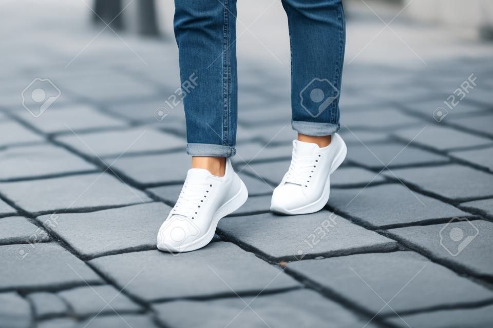 Mulher jovem moderna em um jeans azul elegante em tênis brancos da moda fica em uma estrada de pedra na cidade.