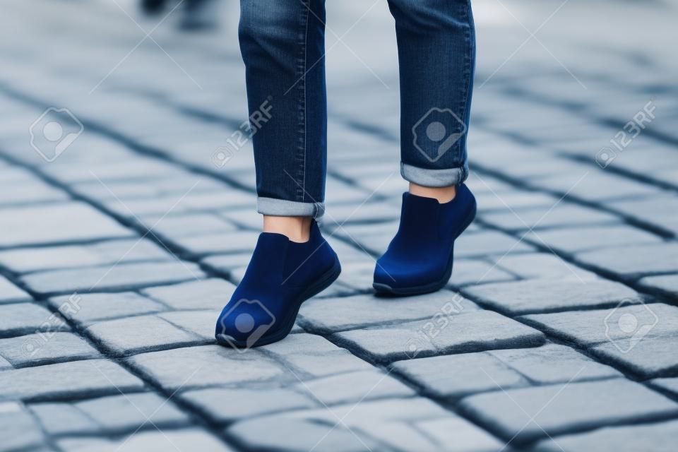 Mulher jovem moderna em um jeans azul elegante em tênis brancos da moda fica em uma estrada de pedra na cidade.