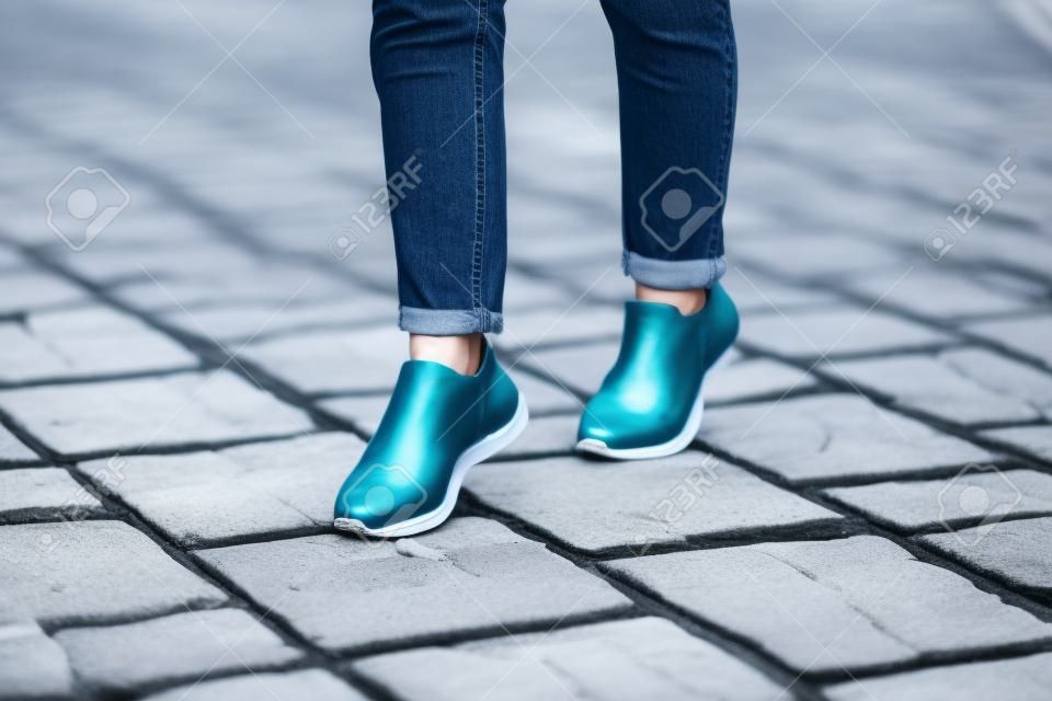 Mujer joven moderna en un elegante jeans azul en zapatillas blancas de moda se encuentra en un camino de piedra en la ciudad.