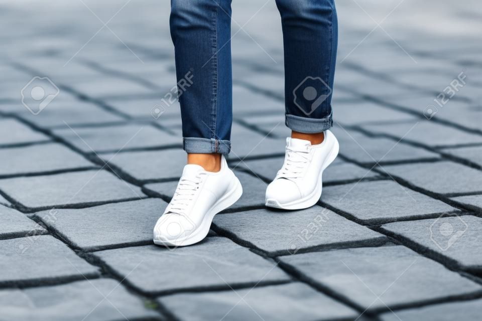La giovane donna moderna in jeans blu alla moda in scarpe da ginnastica bianche alla moda si trova su una strada di pietra della città.