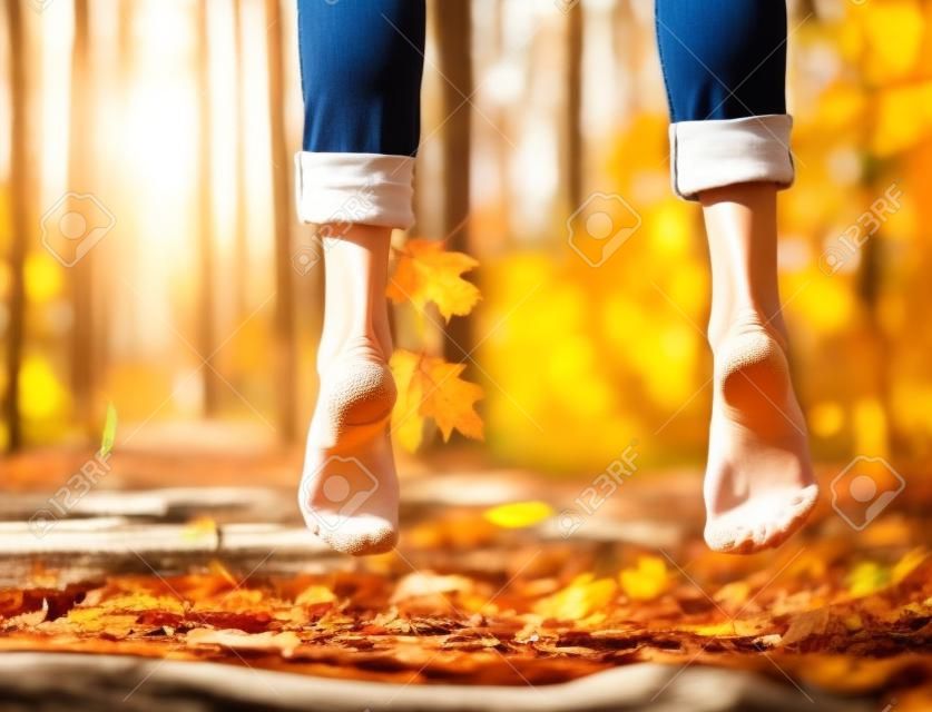 女性健康的腿或脚赤脚和脚跟和脚趾跳在木材上阳光明媚的户外在秋季森林与季节树叶在自然背景为自由的象征。