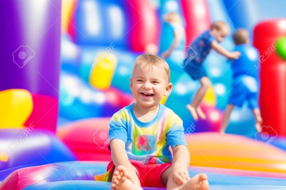 Lächelnd, glücklich barfuß kleiner Junge sitzt auf einem bunten aufblasbaren Plastik Hüpfburg auf einem Rummelplatz oder Spielplatz