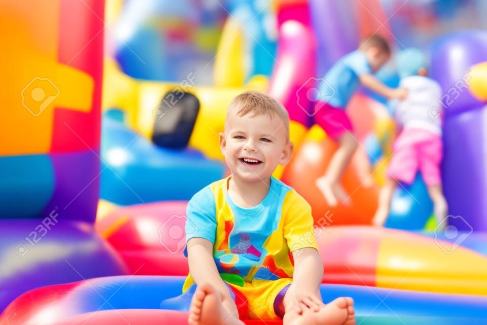 Lächelnd, glücklich barfuß kleiner Junge sitzt auf einem bunten aufblasbaren Plastik Hüpfburg auf einem Rummelplatz oder Spielplatz