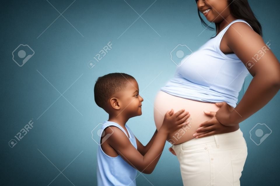 Vue de côté de petit garçon mignon en jeans bleu embrassant le ventre de maman enceinte obscurci avec les mains sur les hanches