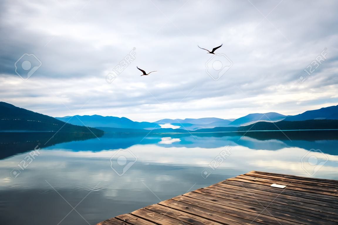 Moody landscape with dark clouds reflecting in calm lake with view of mountains and flying bird in sky