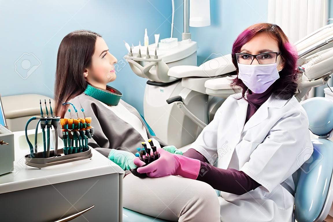 Female dentist with woman patient in the chair at the dental office. Doctor wearing glasses, mask, white uniform and pink gloves. Dentistry. Dental equipment